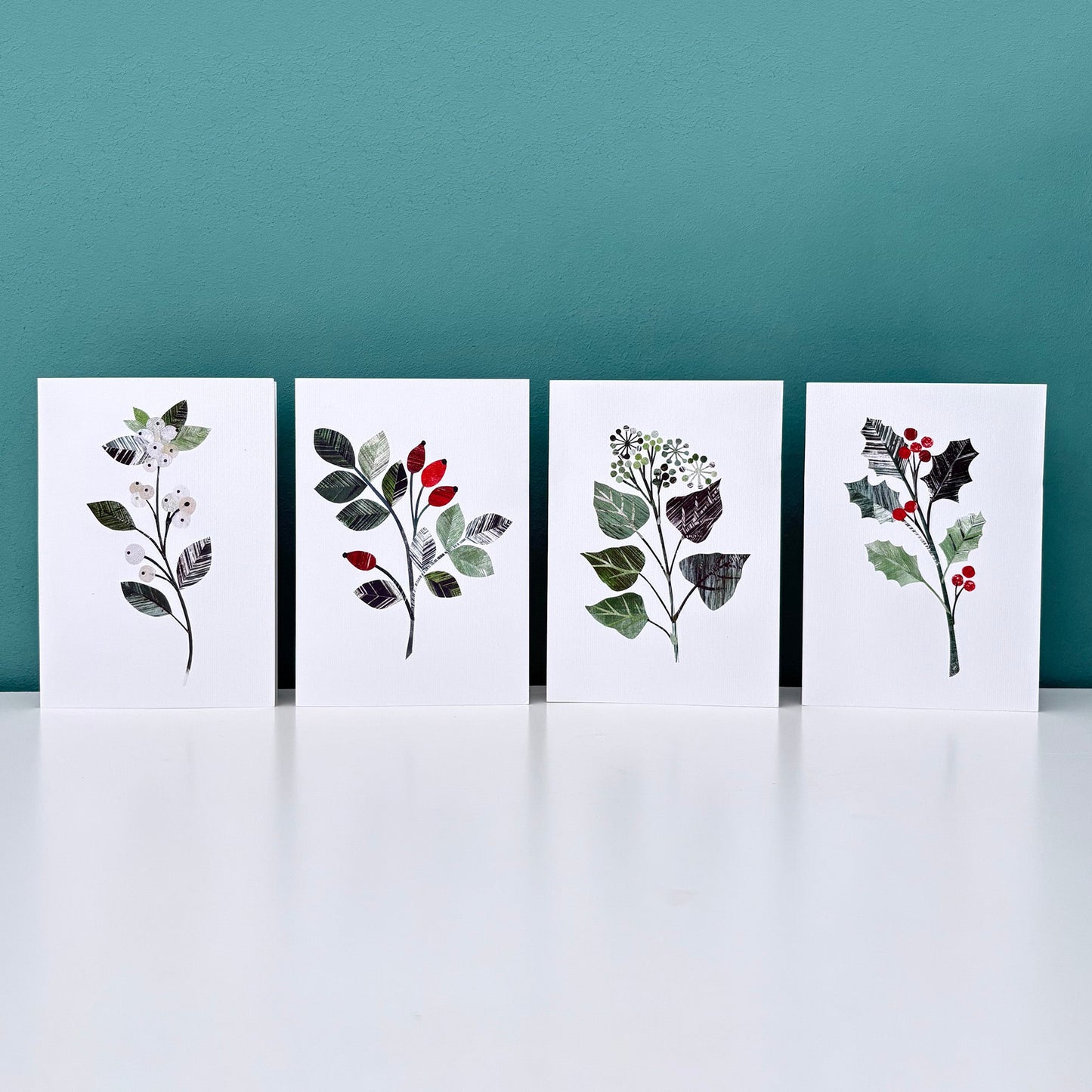 Four Winter floral greeting cards, one with holly, one Rose hips, one Ivy and one Snowberry designs on white backgrounds against a teal wall.