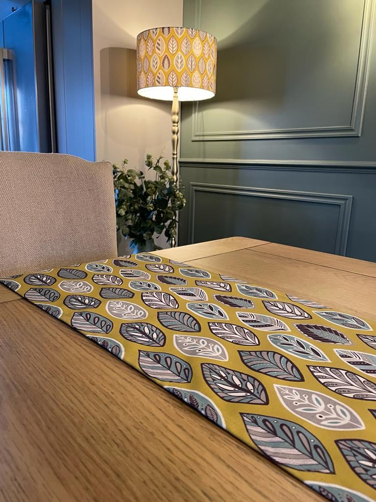 Large Yellow Beech Leaf Lampshade. Displayed here on a Brass floor lamp in the background. A matching yellow beech leaf table runner can be seen on the oak table at the front of this picture. The design features a leaf motif in a Skandinavian style featuring Blue, grey and white leaf shapes on a yellow background.