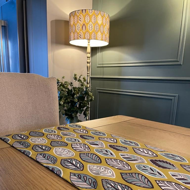 Large Yellow Beech Leaf Lampshade. Displayed here on a Brass floor lamp in the background. A matching yellow beech leaf table runner can be seen on the oak table at the front of this picture. The design features a leaf motif in a Skandinavian style featuring Blue, grey and white leaf shapes on a yellow background.