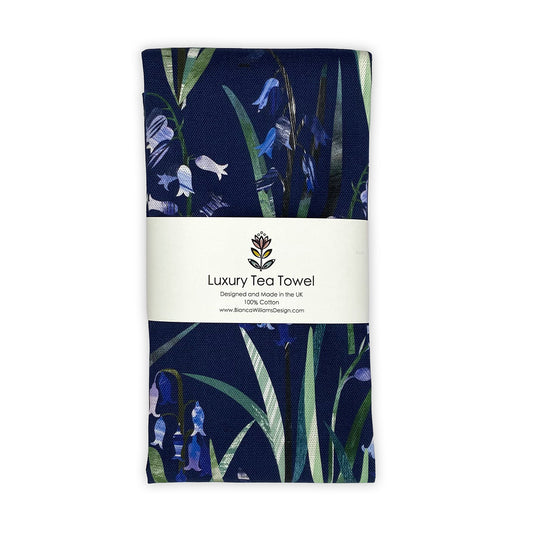 A Navy Blue Bluebell Tea Towel featuring hand collaged Bluebell motifs with the pretty little bluebell flowers and green leaves has been packaged and placed on a plain white background.