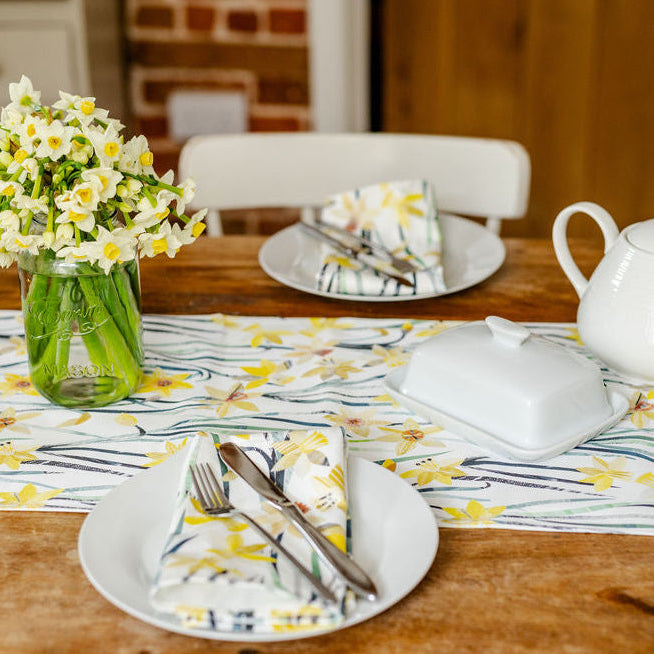 Dining table set with plates, cutlery, a vase of flowers, and a teapot with a Daffodil table runner and matching napkins.