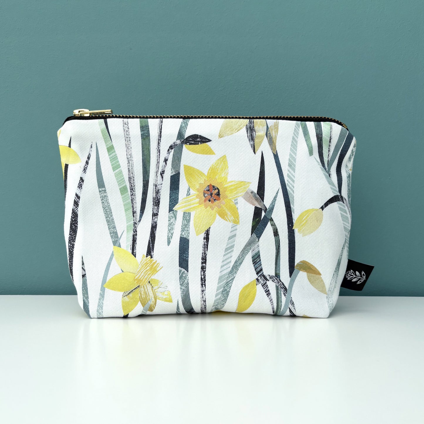 A Daffodil make up bag has been placed on a white shelf against a blue green backdrop.
