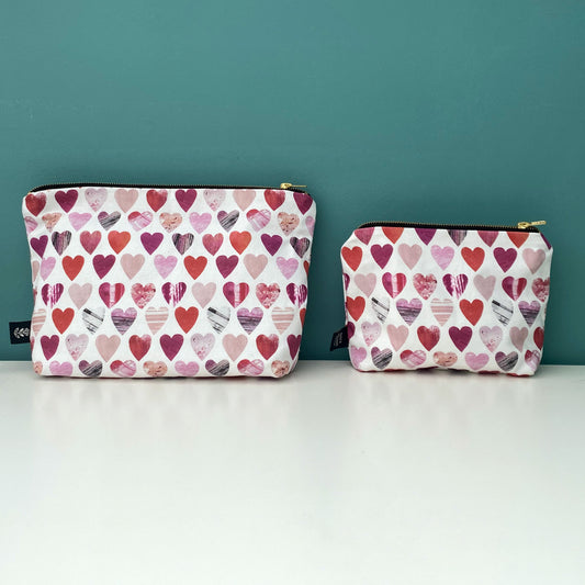 A large and small Hearts Wash Bag have been placed side by side on a white shelf. They have gold metal zip and a Brande label sewn into the outside seam.