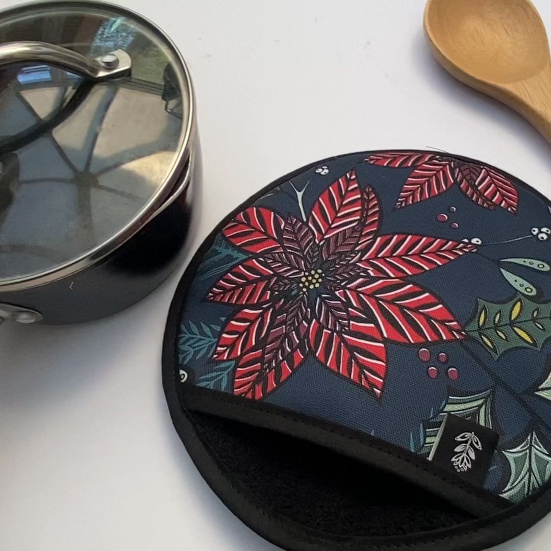 A navy poinsettia pot holder on a white background with a saucepan and wooden spoon.