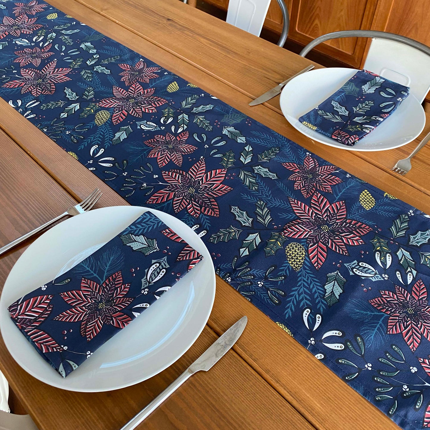 A Navy Poinsettia cotton table runner has been placed down the centre of a wooden table. The table has been laid for two and two matching napkins can also be seen on white plates.