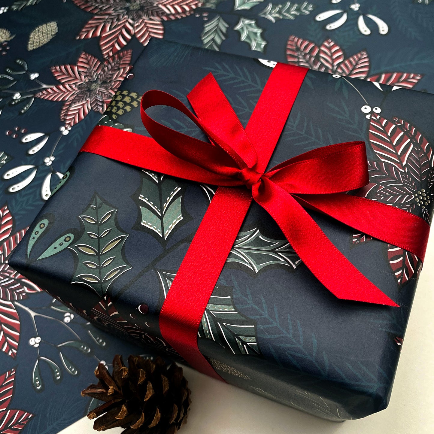A present has been wrapped in the Navy poinsettia gift wrap and it has been tied in a red satin ribbon in a bow. It has been placed on a sheet of gift wrap of the same design and a pine cone has been placed in front.