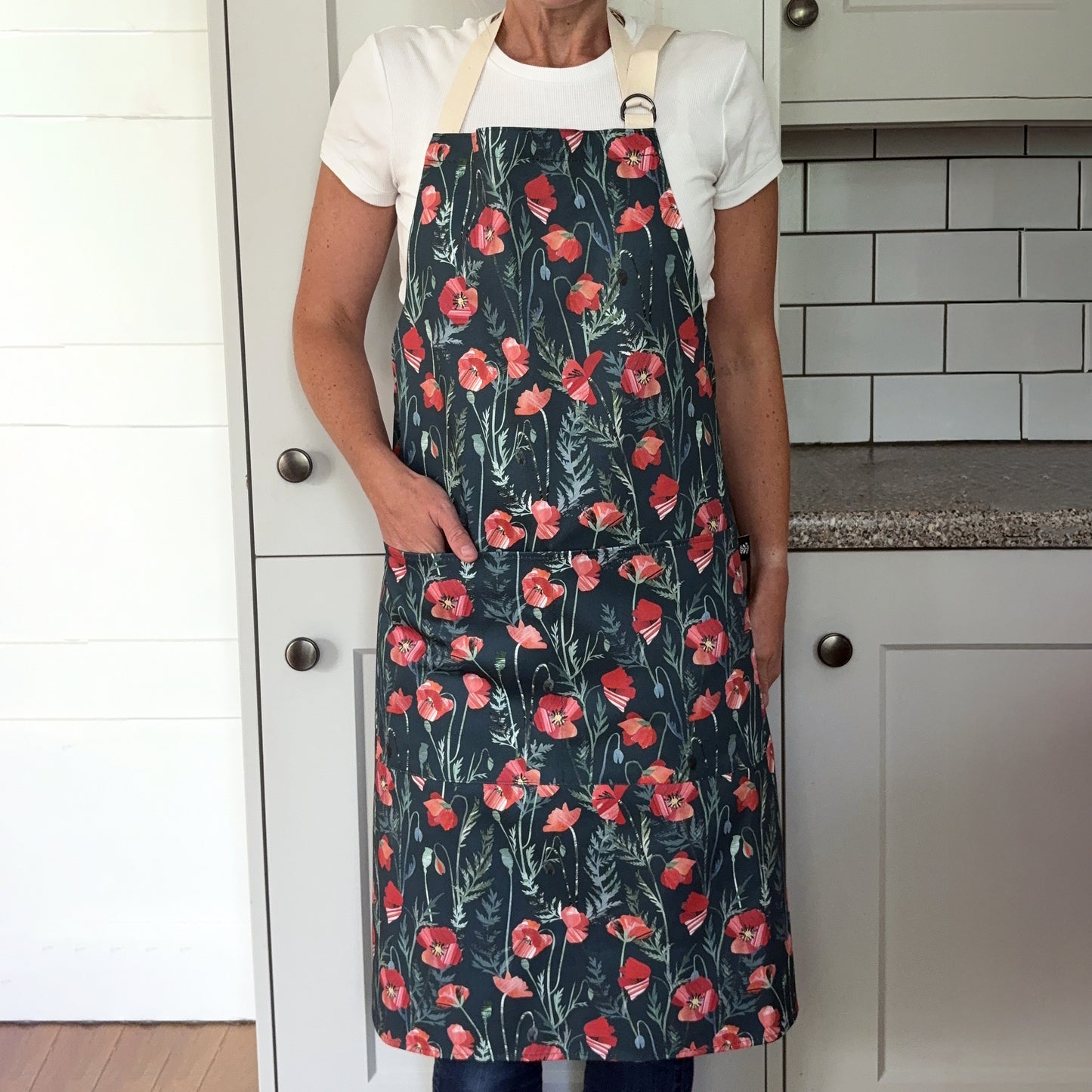 A lady is wearing the Poppy apron with two pockets on the front and an adjustable neck strap in a grey kitchen. She has her hand in one of the front pockets.
