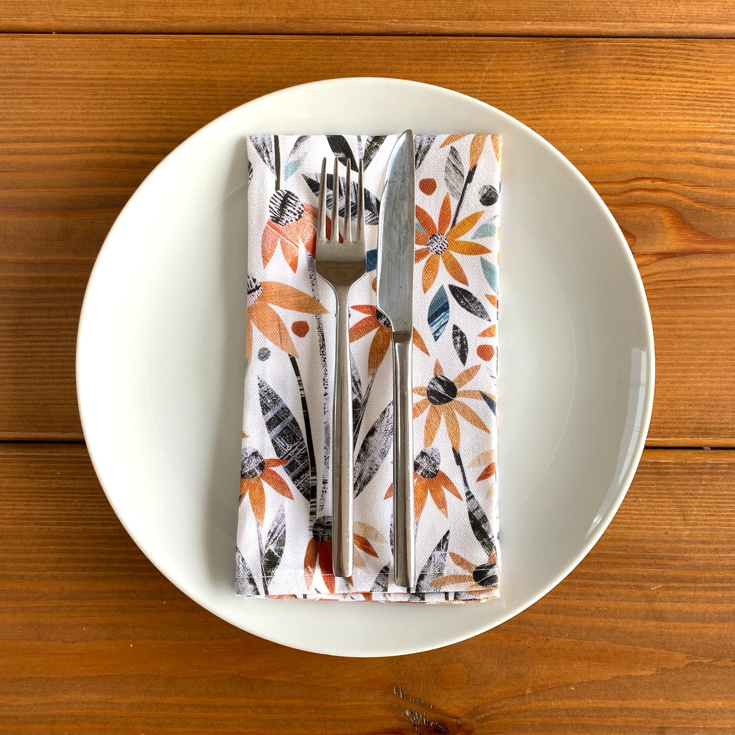 A white plate with an orange Rudbeckia Napkin has been placed on a wooden table.