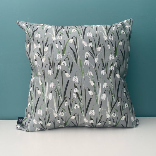 A Snowdrop Cushion featuring pretty little cream and white snowdrop flowers with green leaves on a soft sage green background. The cushion has been placed on a white shelf against a blue/green wall.