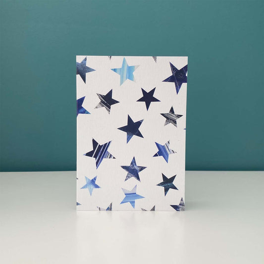 A white textured greeting card with a pattern of textured blue star cutouts, standing upright on a white surface against a teal background.