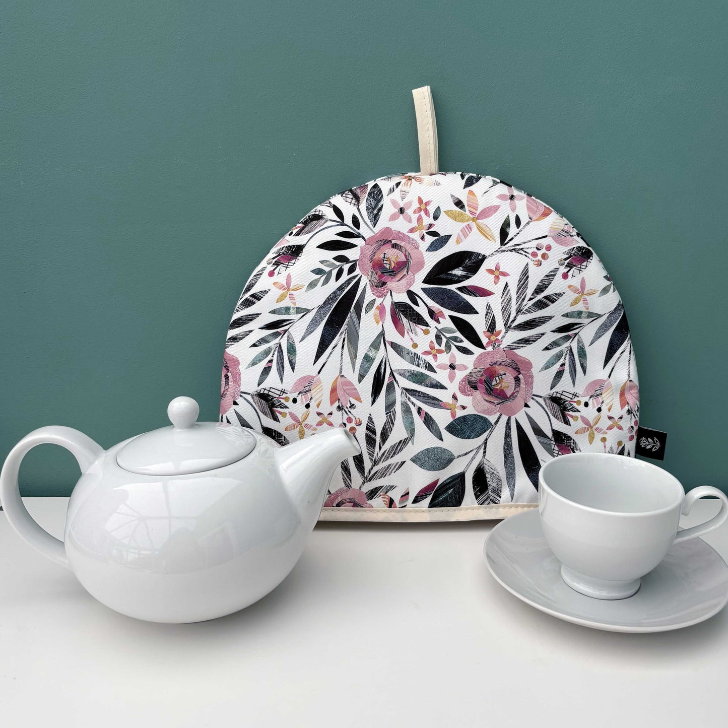 A Summer Floral Tea Cosy has been placed on a white shelf with a white tea pot, tea cup and saucer against a teal wall.