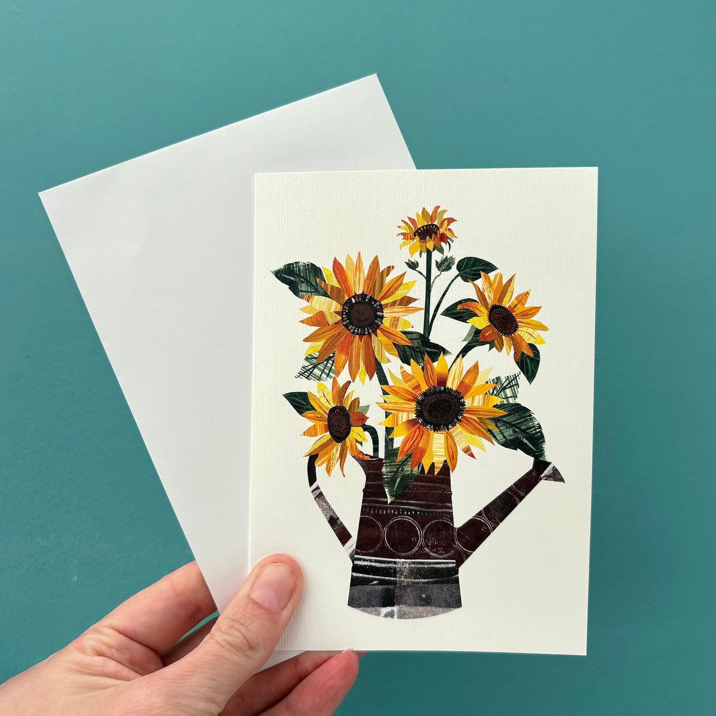 A white card with an image of yellow and Orange sunflowers with green leaves in a watering can, is being held with a white envelope against a blue green background.