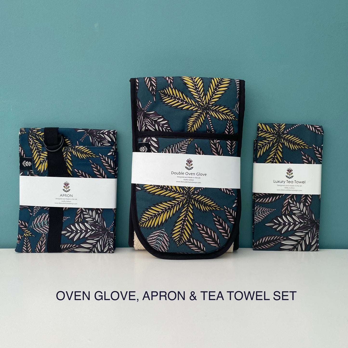 A packaged set which includes a matching Teal Forest Floor Apron, Double Oven Gloves and Tea Towel. They have been placed on a white shelf with a blue/green background.
