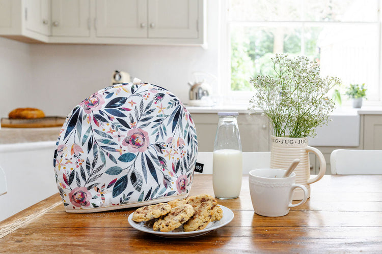 A Summer Floral Tea Cosy has been placed on a wooden table in a kitchen with a small plate of cookies, a cup of tea and a vase of flowers.