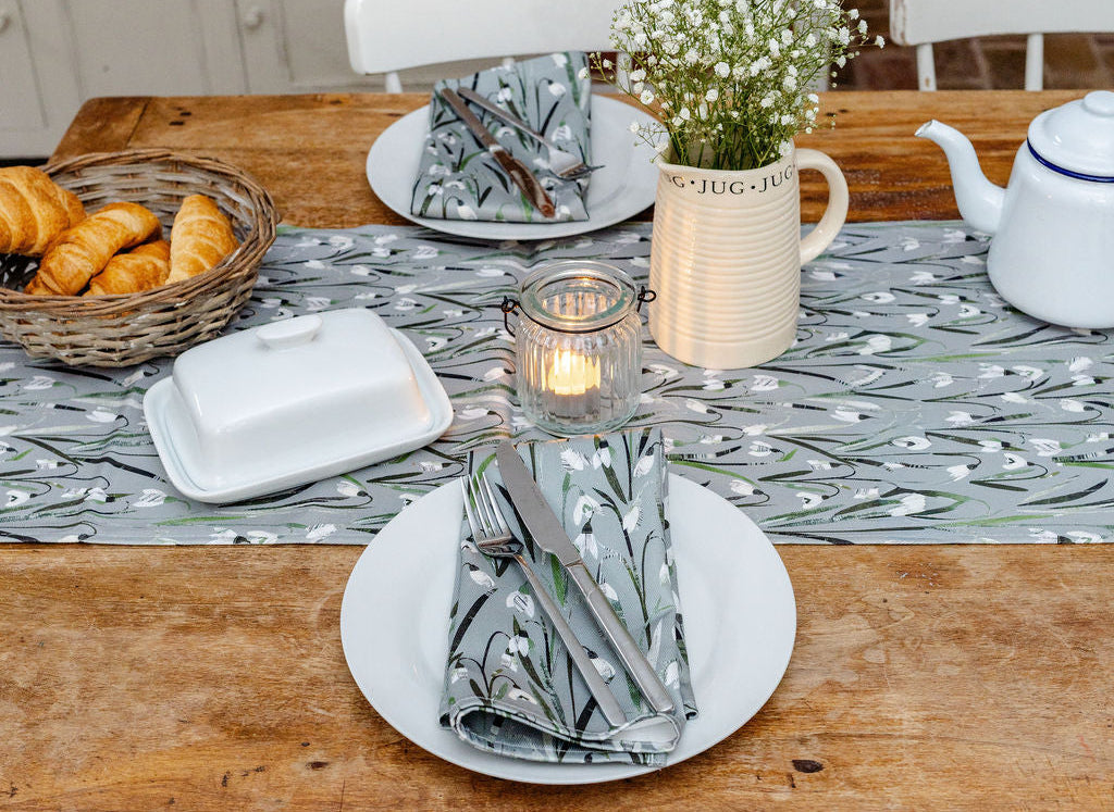 A rustic dining table set for two, featuring a gray and white fish-patterned table runner and matching napkins, white plates, a small candle, a ceramic vase with baby's breath, and a white teapot on a weathered wooden table.