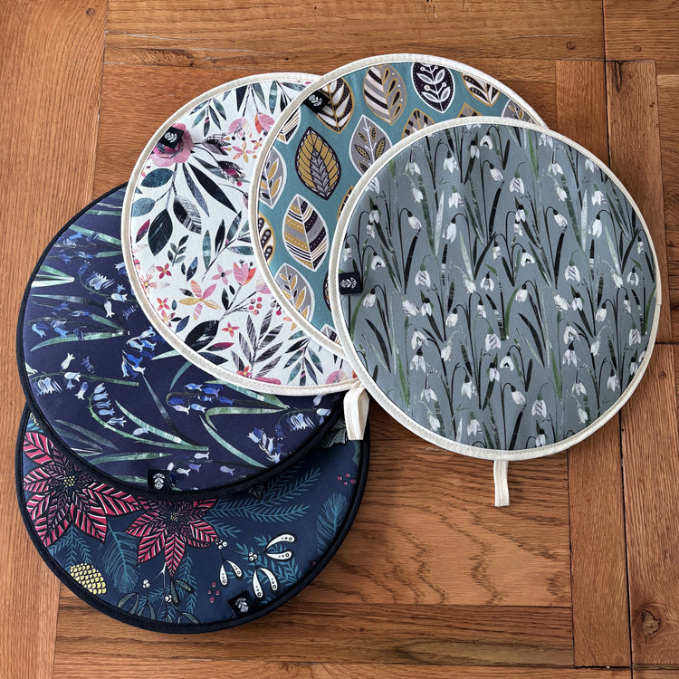 A selection of hob covers suitable for use with AGA have been placed on a wooden table.  Designs include one navy Poinsettia Christmas Hove Cover, a Navy Bluebells, Summer Floral, Snowdrops and Blue Beech Leaf Circular hob covers