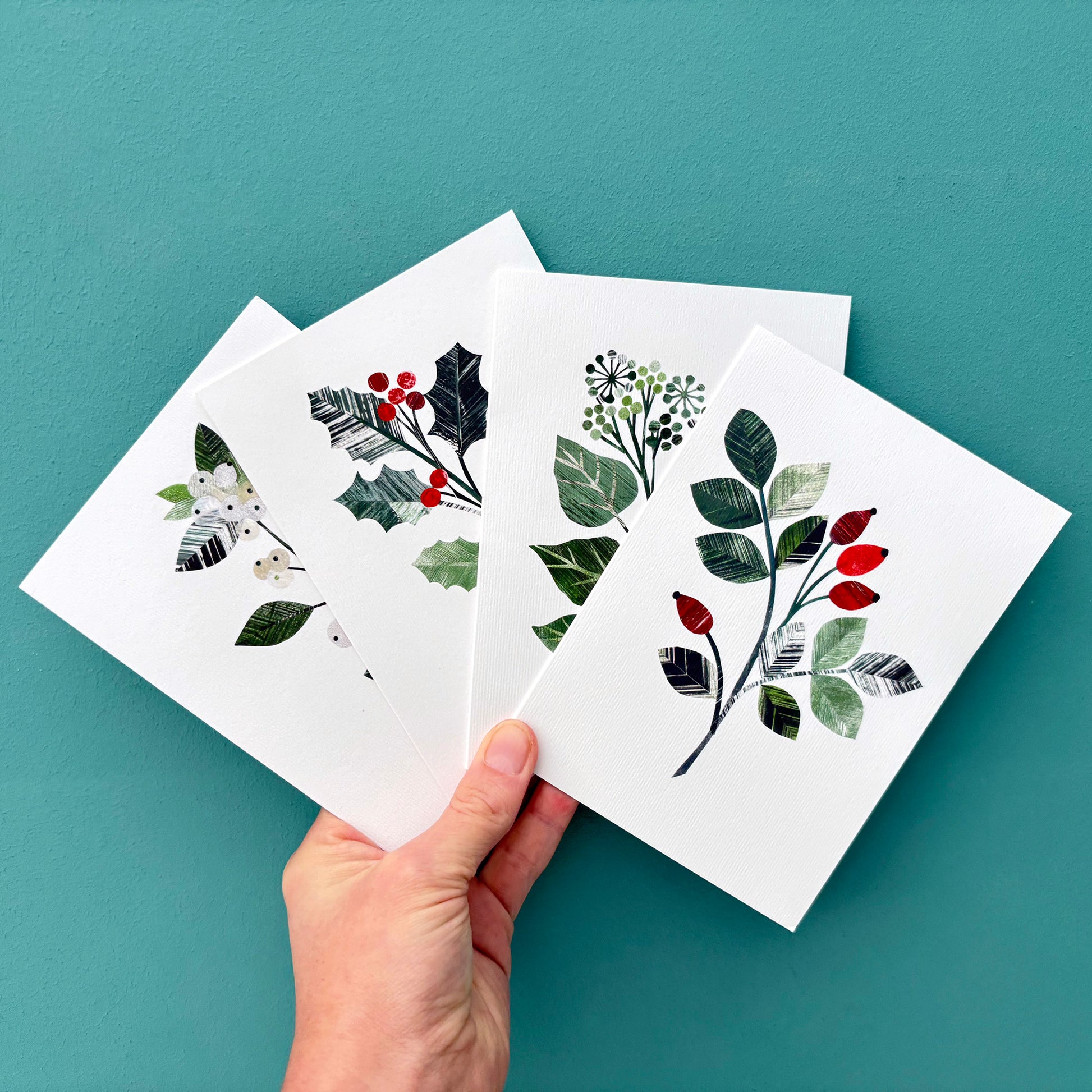 Hand holding four white cards with winter foliage designs on a teal background.