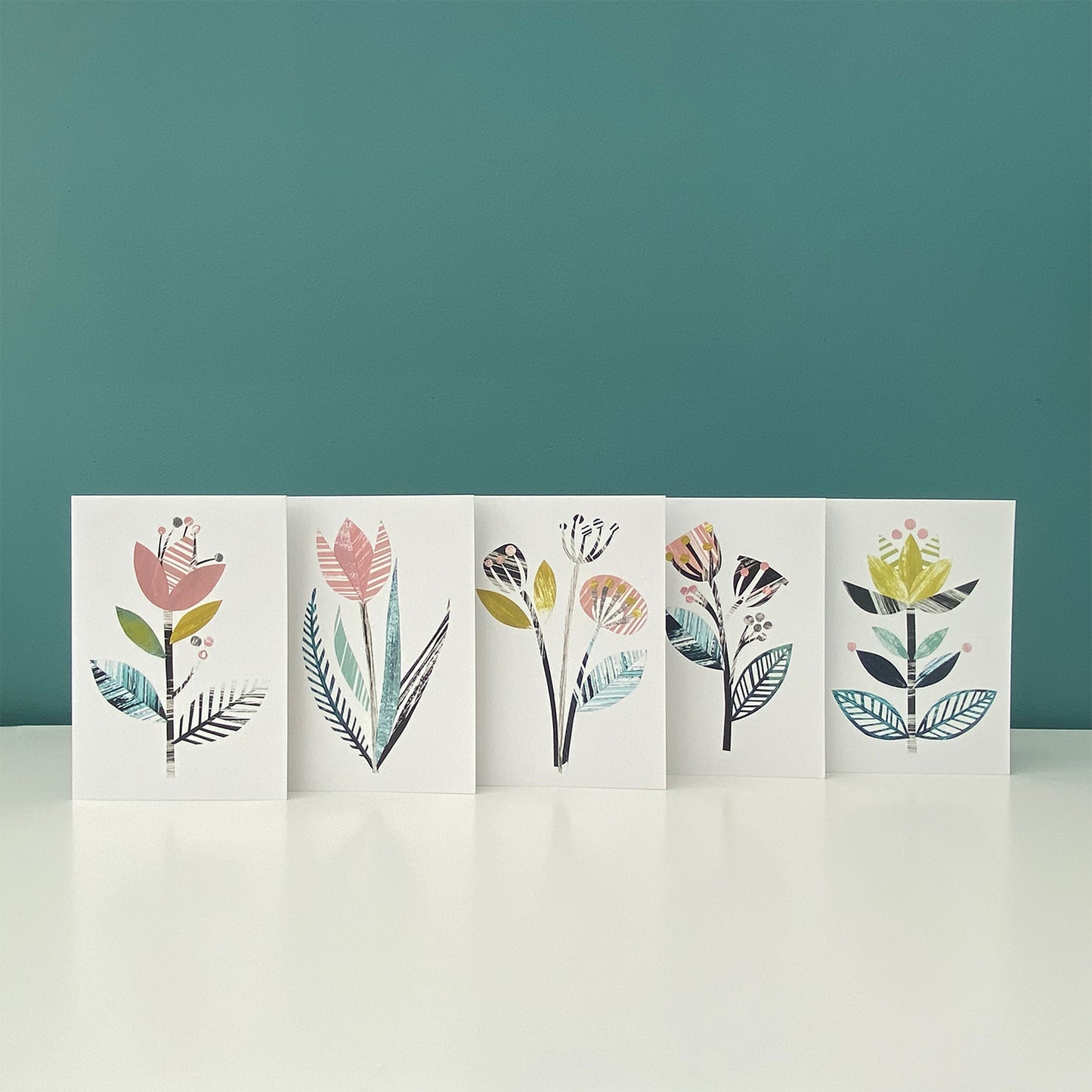 Five different but coordinating stylised flower cards in the same colour palette of yellow, green, dusky pink and greys have been stood together in a row on a white shelf with a blue green. 