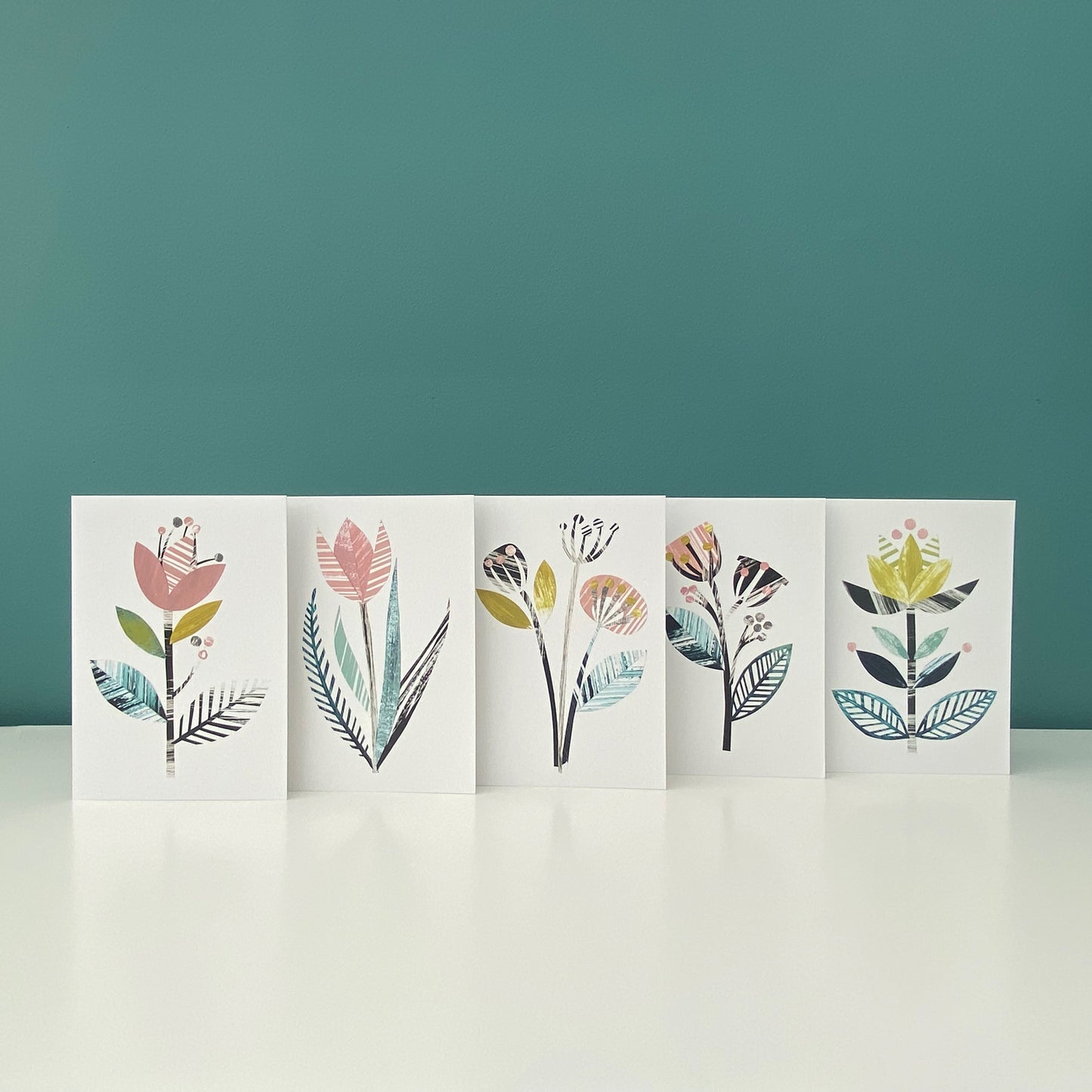 Five different but coordinating stylised flower cards in the same colour palette of yellow, green, dusky pink and greys have been stood together in a row on a white shelf with a blue green background.