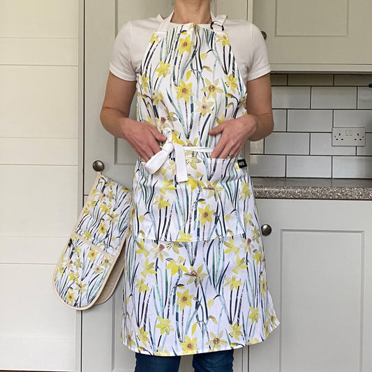 Lady wears the Daffodil Apron featuring the pretty yellow and green flowers on a white background.  The Apron has two pockets and an adjustable neck strap.  