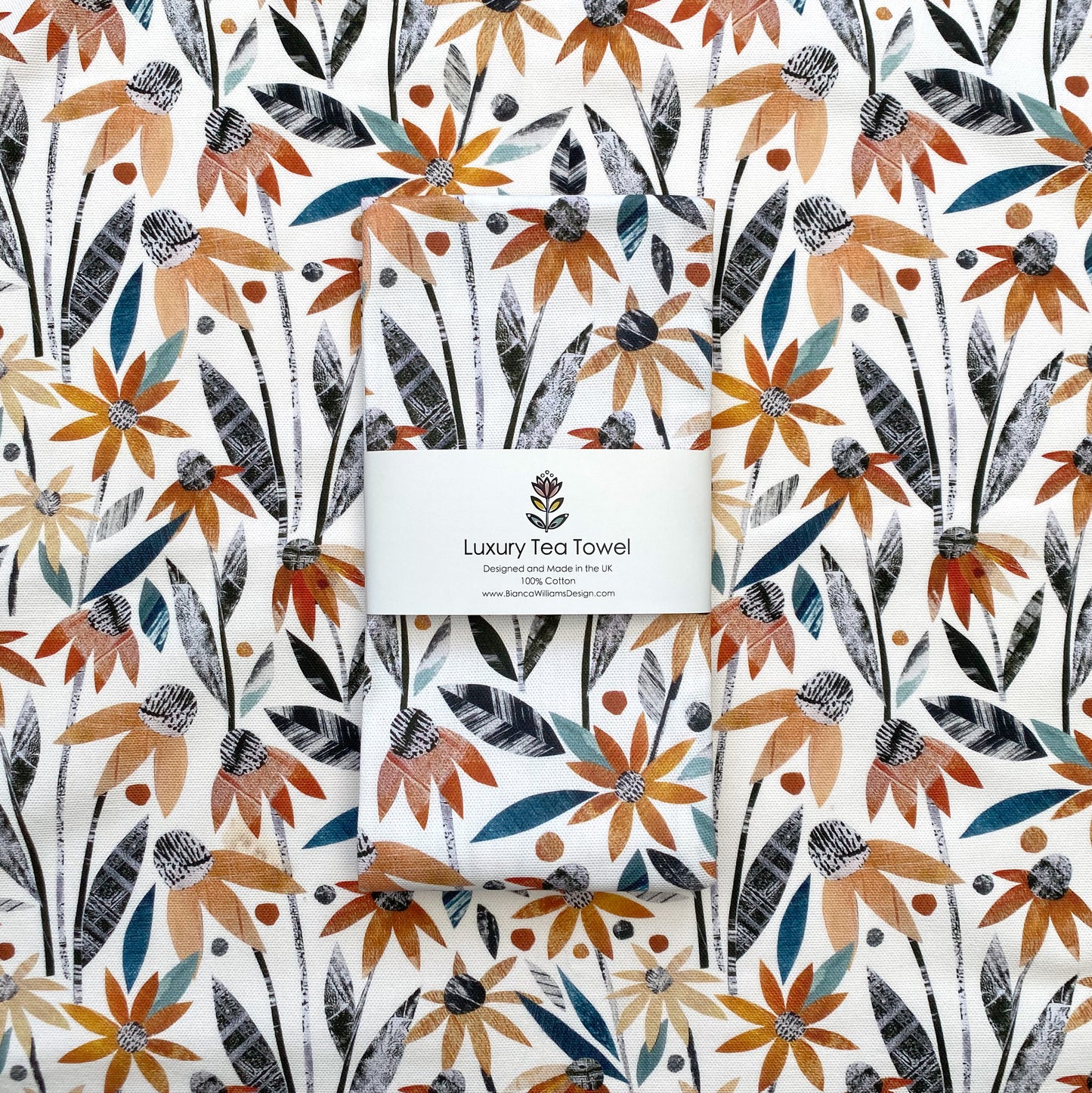 Rudbeckia Cotton Tea Towel, featuring Coneflowers in every shade of orange with Soft Green and Grey Leaves on a white background.  One Rudbeckia Tea Towel is shown in packaging laid onto a laid out Rudbeckia tea Towel.