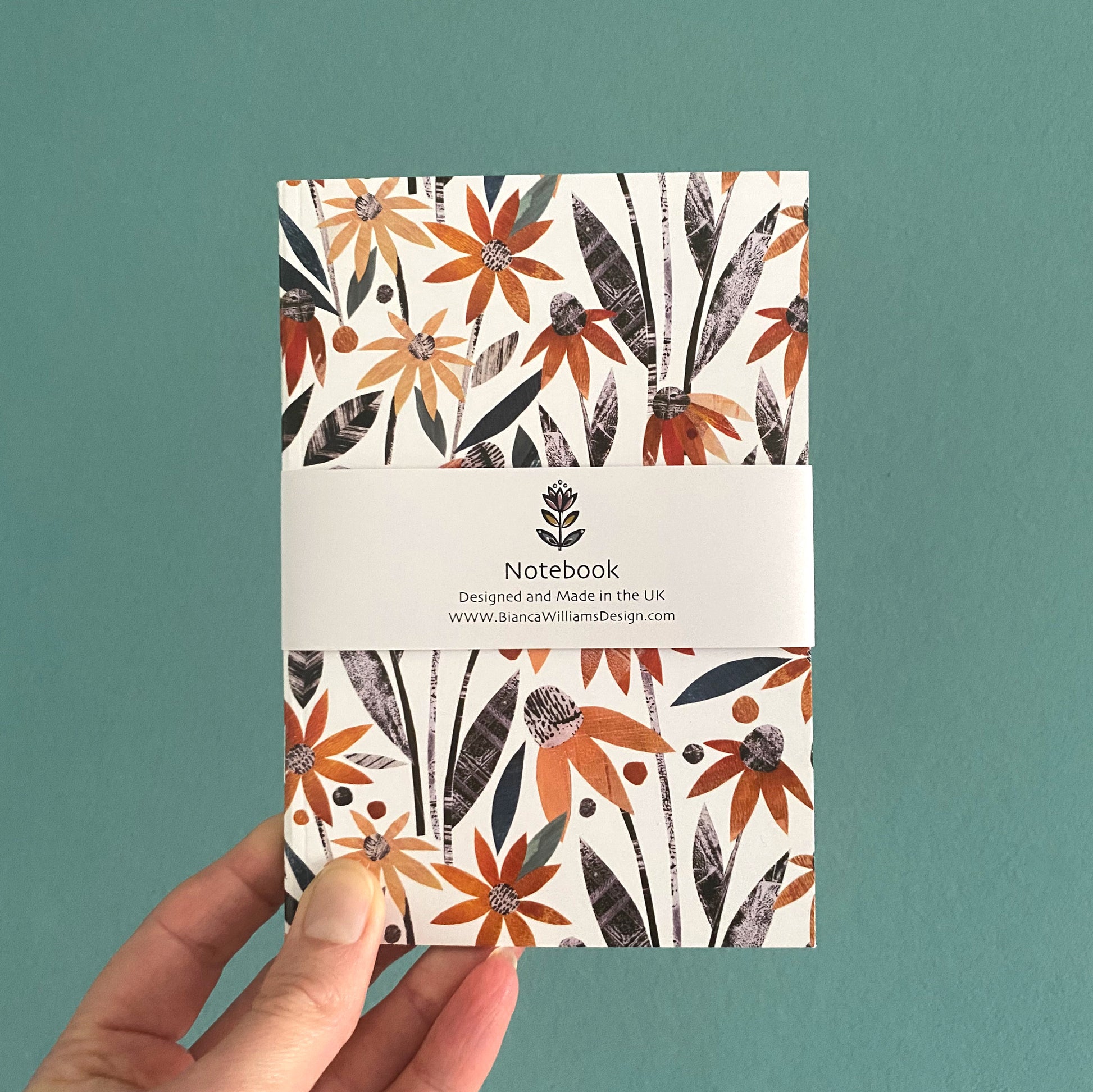 Small A6 notebook featuring orange all over Coneflower patterns with green and grey leaves on a white background, packaged in a white branded belly band held in front of a blue green backdrop.