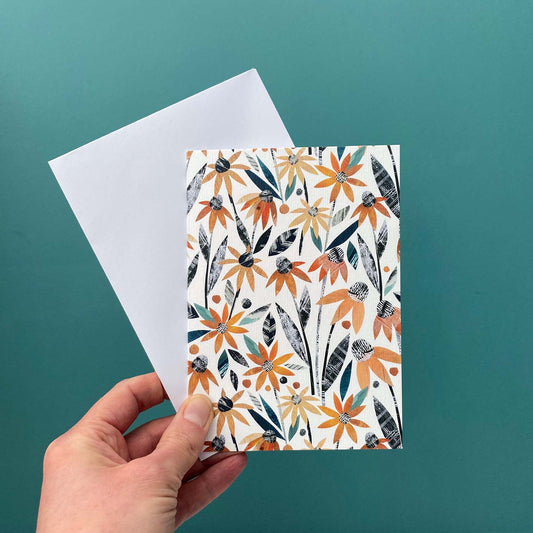 An Autumn Floral Greeting Card featuring Orange rudbeckia all over print on a white background is being held with a white envelope against a blue green wall.