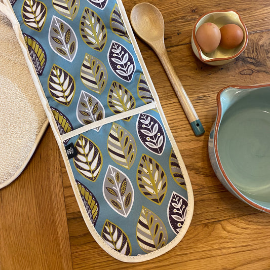 Blue Beech Leaf Oven Gloves, shown laid at an angle on an oak table, so that you can see the Ecru cotton towelling on the reverse.  There is also a wooden spoon and two bowls on the table.