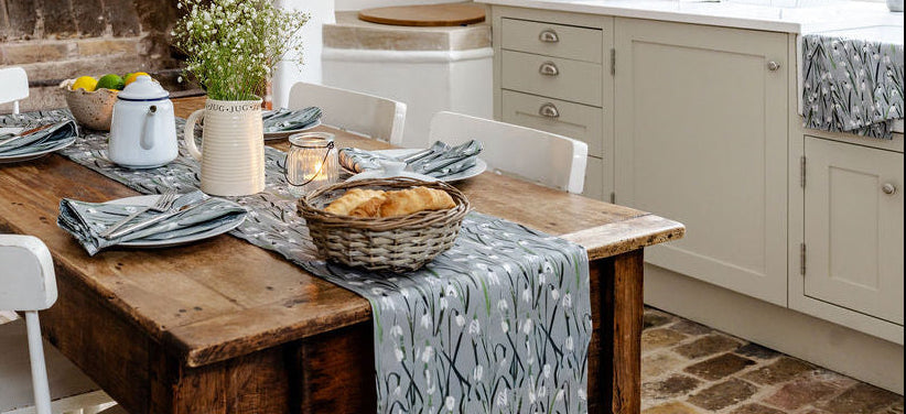 A rustic wooden dining table set with plates, a patterned runner, and a basket of bread, set in a bright kitchen with white cabinets and a brick fireplace.