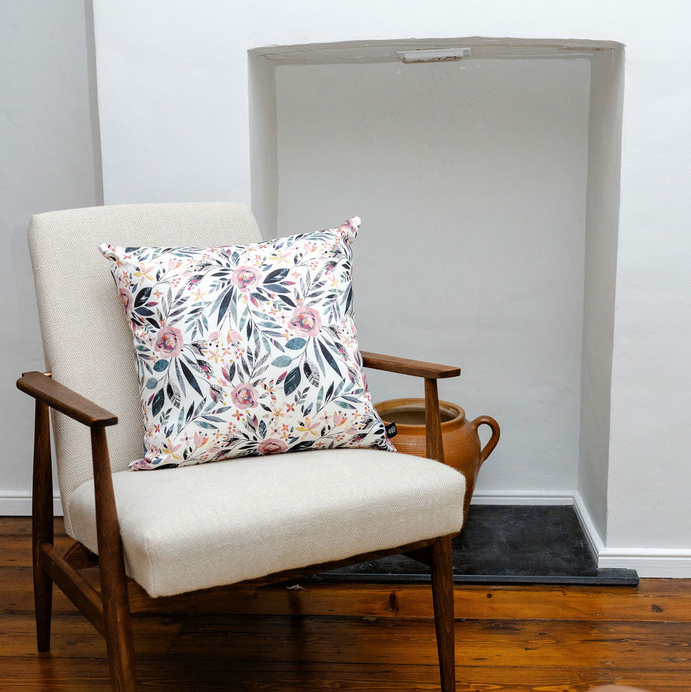 A mid-century modern armchair with cream upholstery and dark wood arms, styled with a square floral throw pillow, set in a room with a fireplace and hardwood floor.