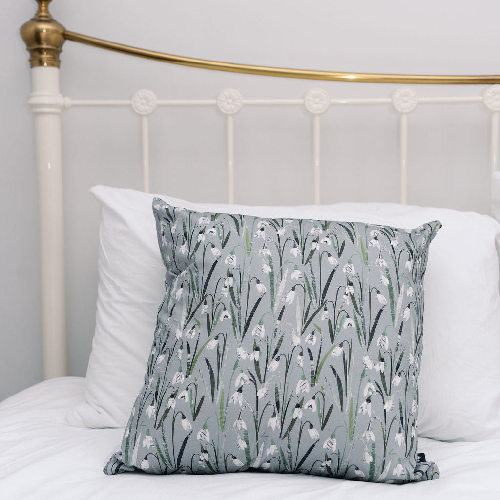 A square decorative throw pillow with a grey and white Snowdrop floral pattern, styled on a white bed with white pillows and a white metal headboard with gold accents.