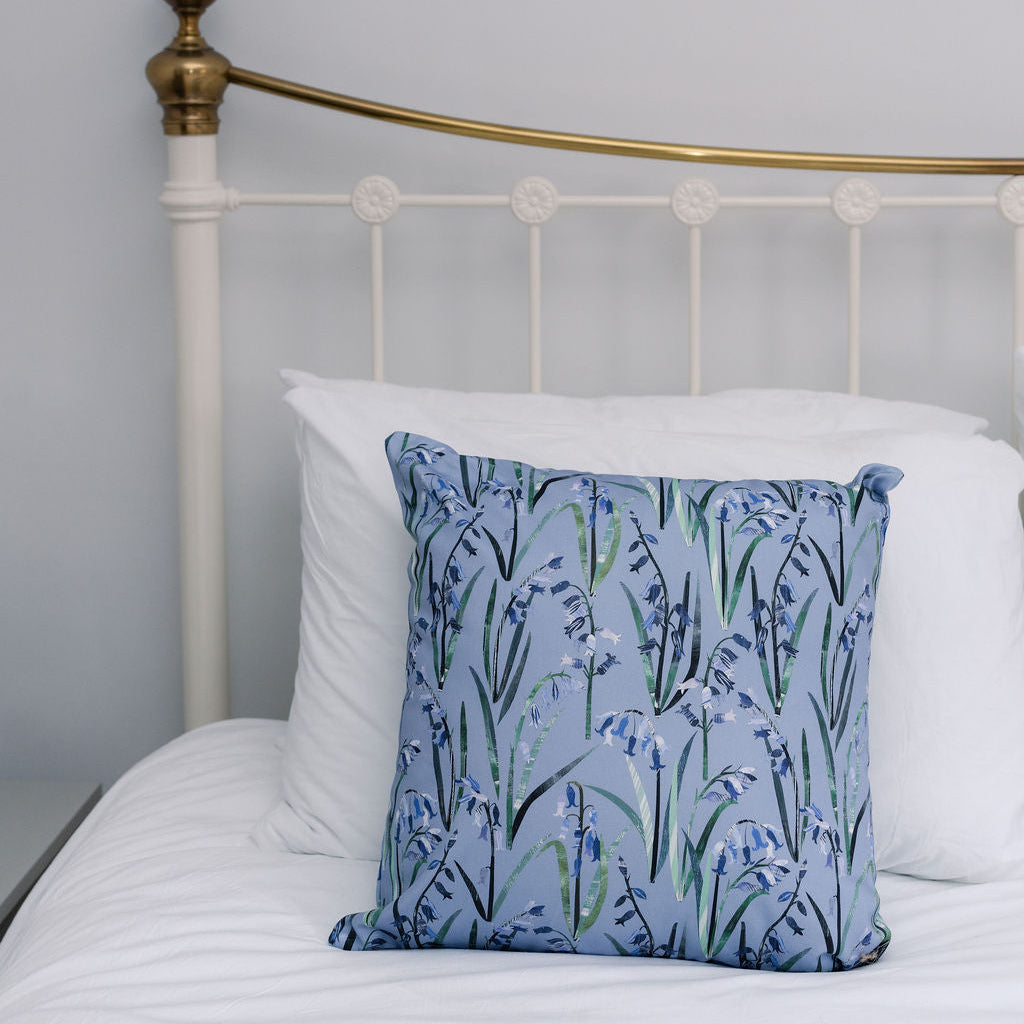 A square decorative throw pillow with a blue and green bluebell floral pattern, styled on a bed with white pillows and a white metal headboard.