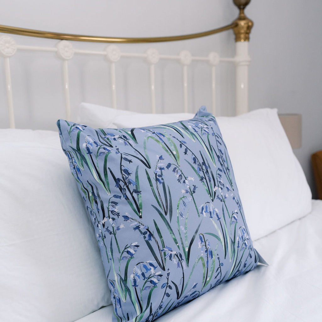 A square decorative throw pillow with a blue and green bluebell floral pattern, styled on a white bed with white pillows in front of a white metal headboard with gold accents.