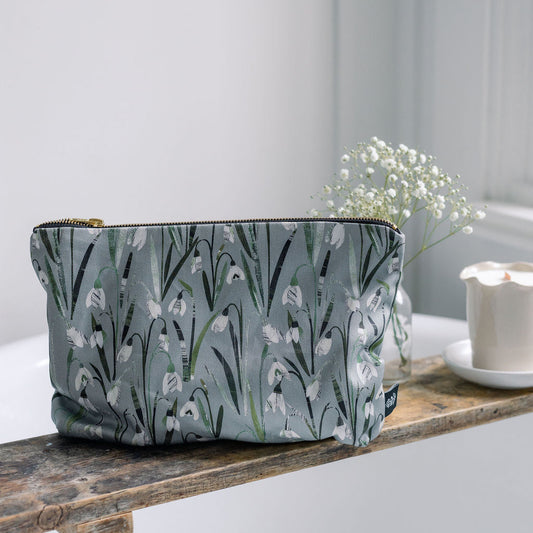 A rectangular zip pouch with a muted green-gray background and an allover white and green floral print, resting on a rustic wooden shelf next to baby's breath flowers and a candle.