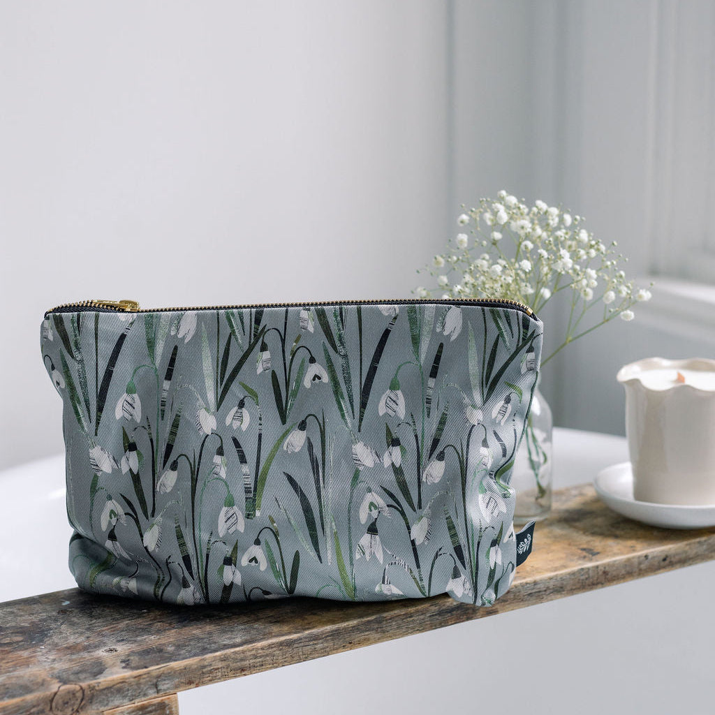 A rectangular zip pouch with a muted green grey background and an allover white and green Snowdrop floral print, resting on a rustic wooden shelf next to baby's breath flowers and a candle.