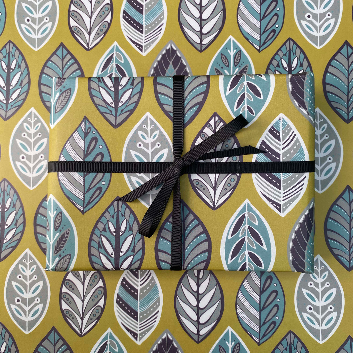 A gift box wrapped in mustard yellow wrapping paper with a repeating pattern of stylized teal, gray, and white leaves, tied with a black ribbon and bow.