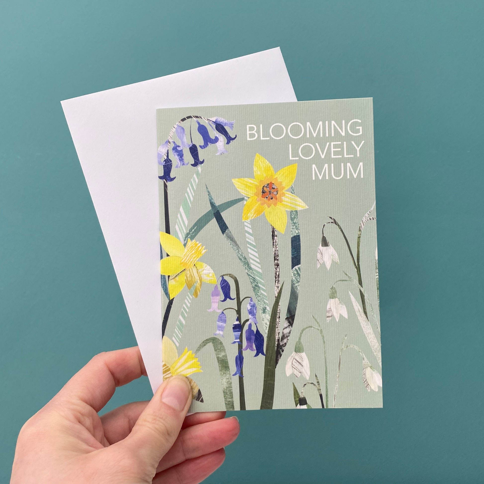 The Blooming Lovely Mum Greeting card is being held with a white envelope against a blue green background.