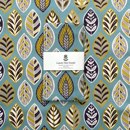 Blue Beech leaf Tea Towel featuring simple leaf motifs in Blue, Yellow, white, grey and aubergine.  A packaged Tea Towel has been placed on an open out tea towel of the same design.