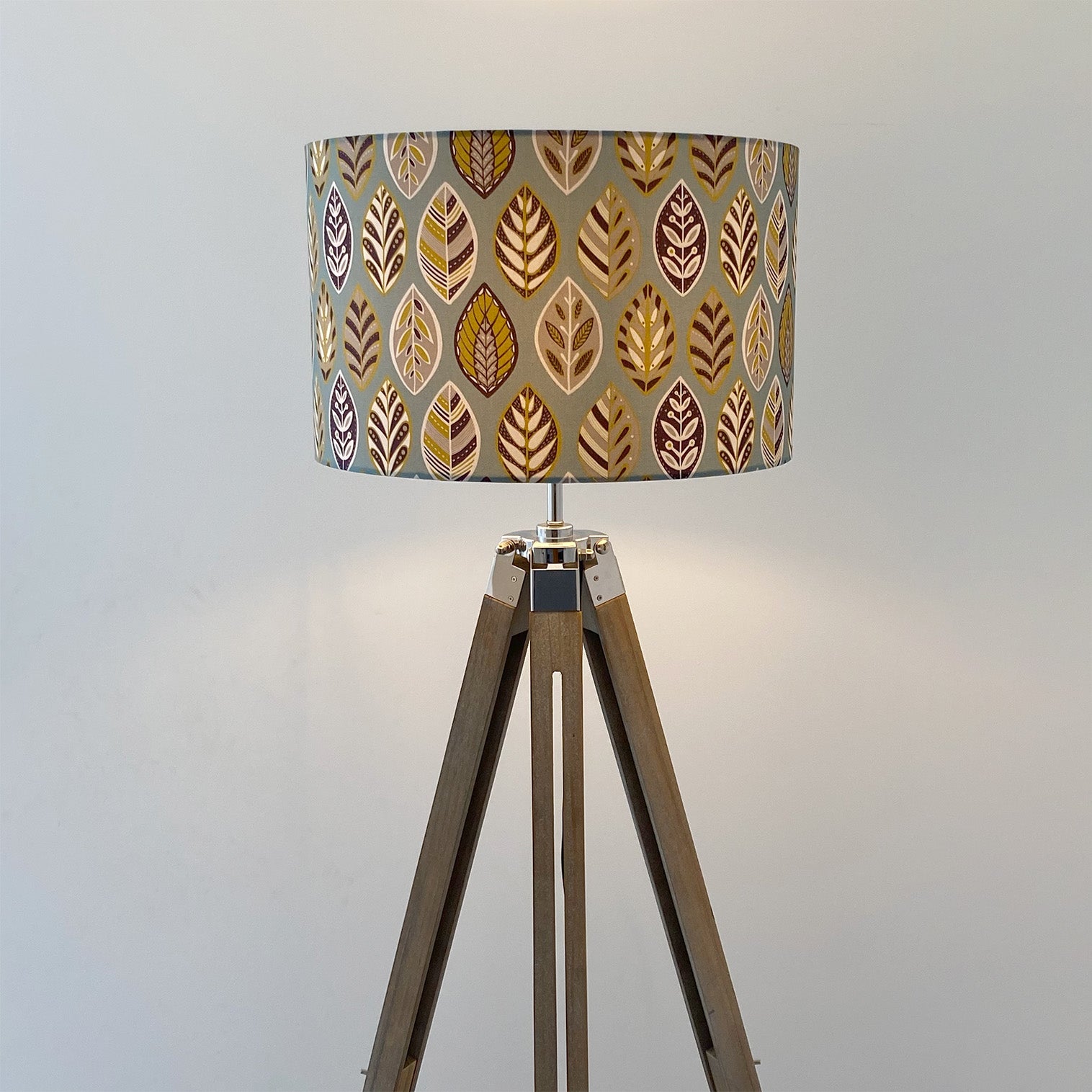 Large Blue Beech leaf Lampshade, shown here on a wooden tripod floor lamp. The pattern features a Skandi style Blue, yellow, grey and white Leaf pattern on a Blue Background.  The light is shown on in this photograph.