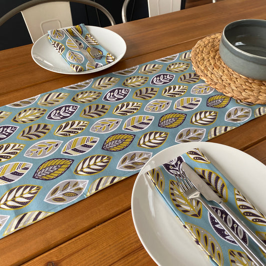 Blue beech leaf table runner placed in the middle of a wooden table.  The table has been set for two and has two white plates on it with matching napkins.