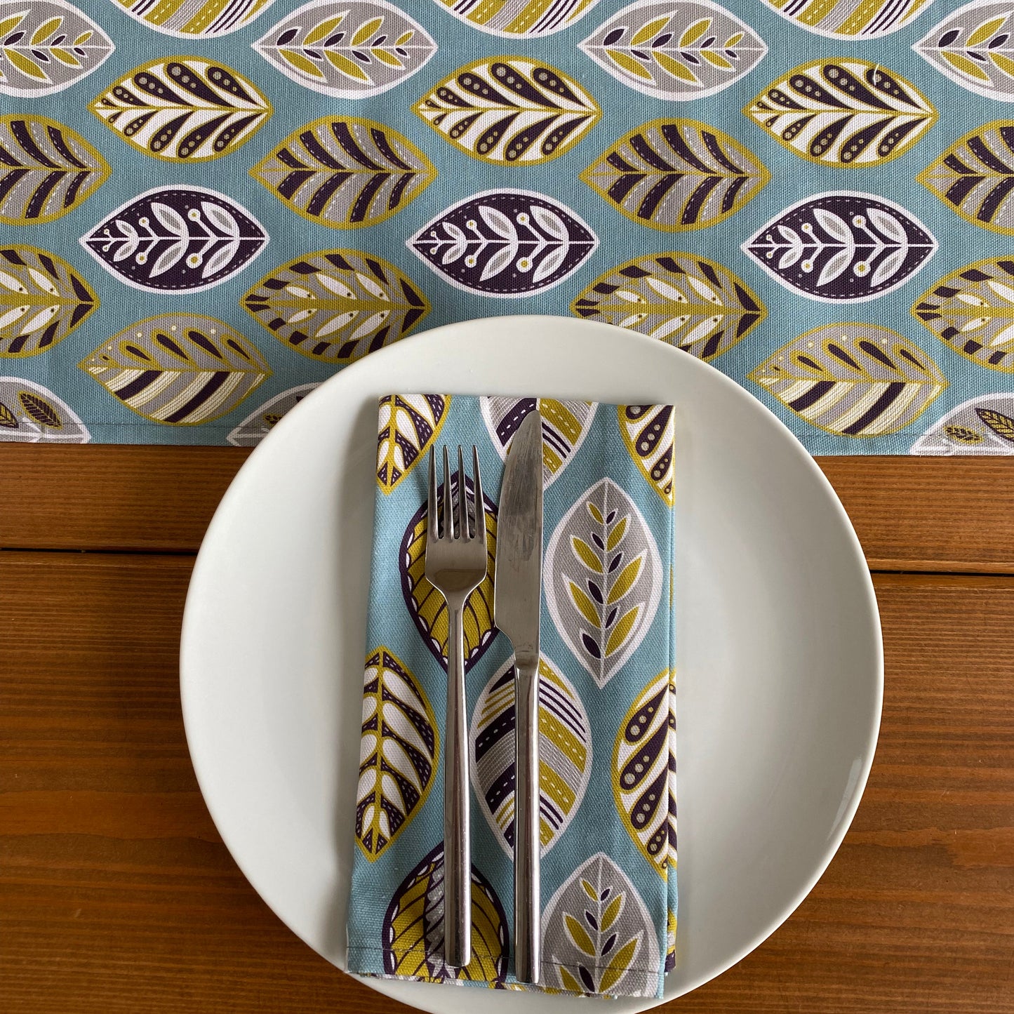 This is a Birds Eye view of the blue beech leaf table runner in the centre of a wooden table.  The table also has a white plate on it with a matching blue beech leaf napkin placed on it.