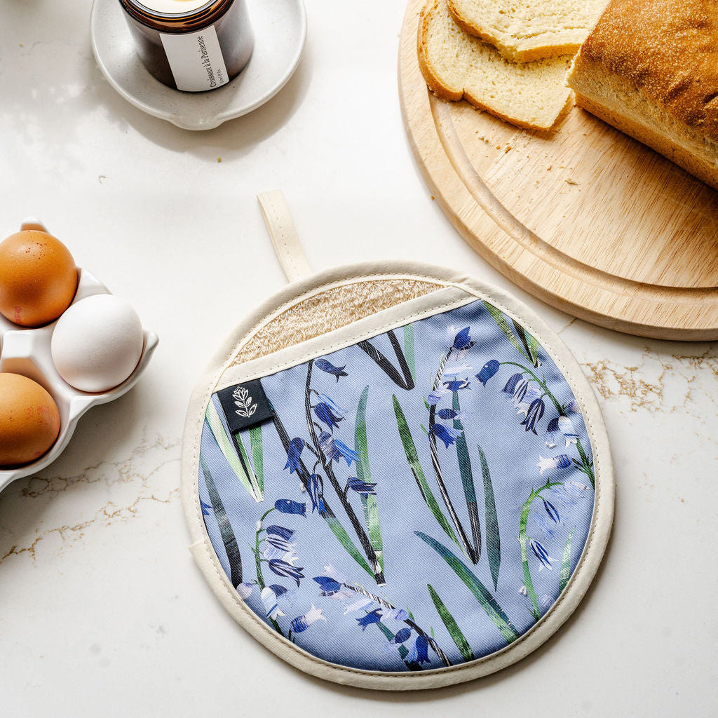 A round potholder with a blue and green bluebell floral print is displayed on a white marble surface, styled with a lit candle, sliced bread on a wooden board, and an egg carton.