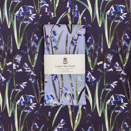 A light blue tea towel featuring Bluebells is folded and packaged in a branded belly band and has been placed on top of an opened out Navy Bluebell Tea Towel featuring the same design.