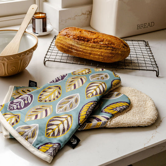 A pair of patterned Long Oven Gloves featuring a stylised leaf pattern on a blue background.  They have beige cotton towelling backing and have been placed on a marble kitchen work top with a loaf of bread and a mixing bowl.