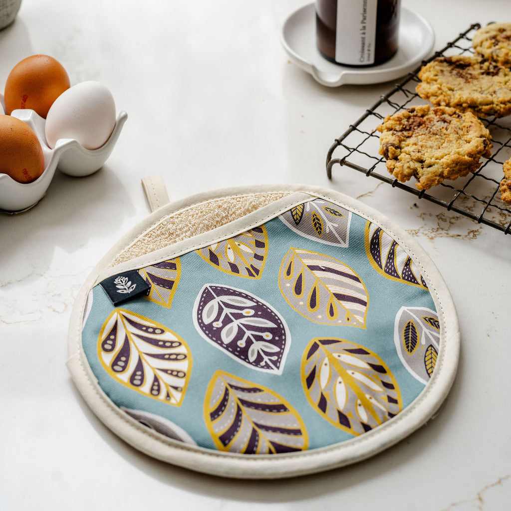 A round potholder with a light blue background and a pattern of stylized leaves in yellow, purple, and white, styled on a white marble kitchen counter next to eggs, cookies, and a candle.