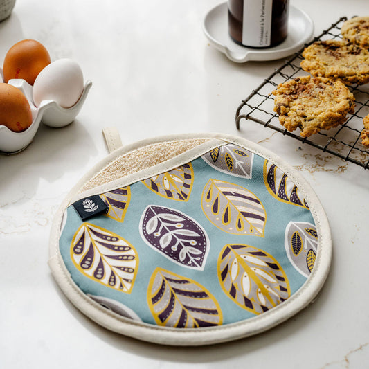 A round potholder with a light blue background and a pattern of stylized leaves in yellow, purple, and white, styled on a white marble kitchen counter next to eggs, cookies, and a candle.