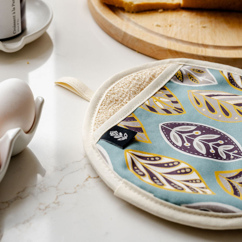 A round potholder with a light blue background and a botanical leaf pattern in purple, yellow, and beige, resting on a white marble countertop next to a bread slice and a white ceramic egg cup.