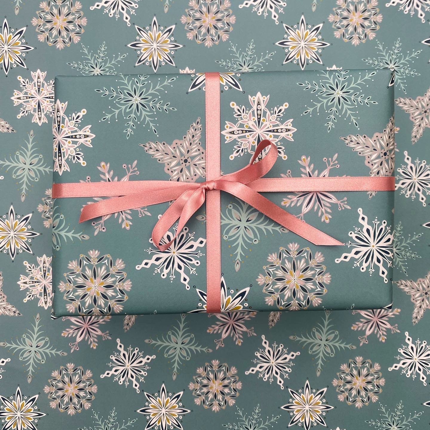 A present has been wrapped in the Blue Snowflake gift wrap and has been tied with coordinating pink satin ribbon in a bow.  It has been placed on a sheet of the blue snowflake gift wrap.