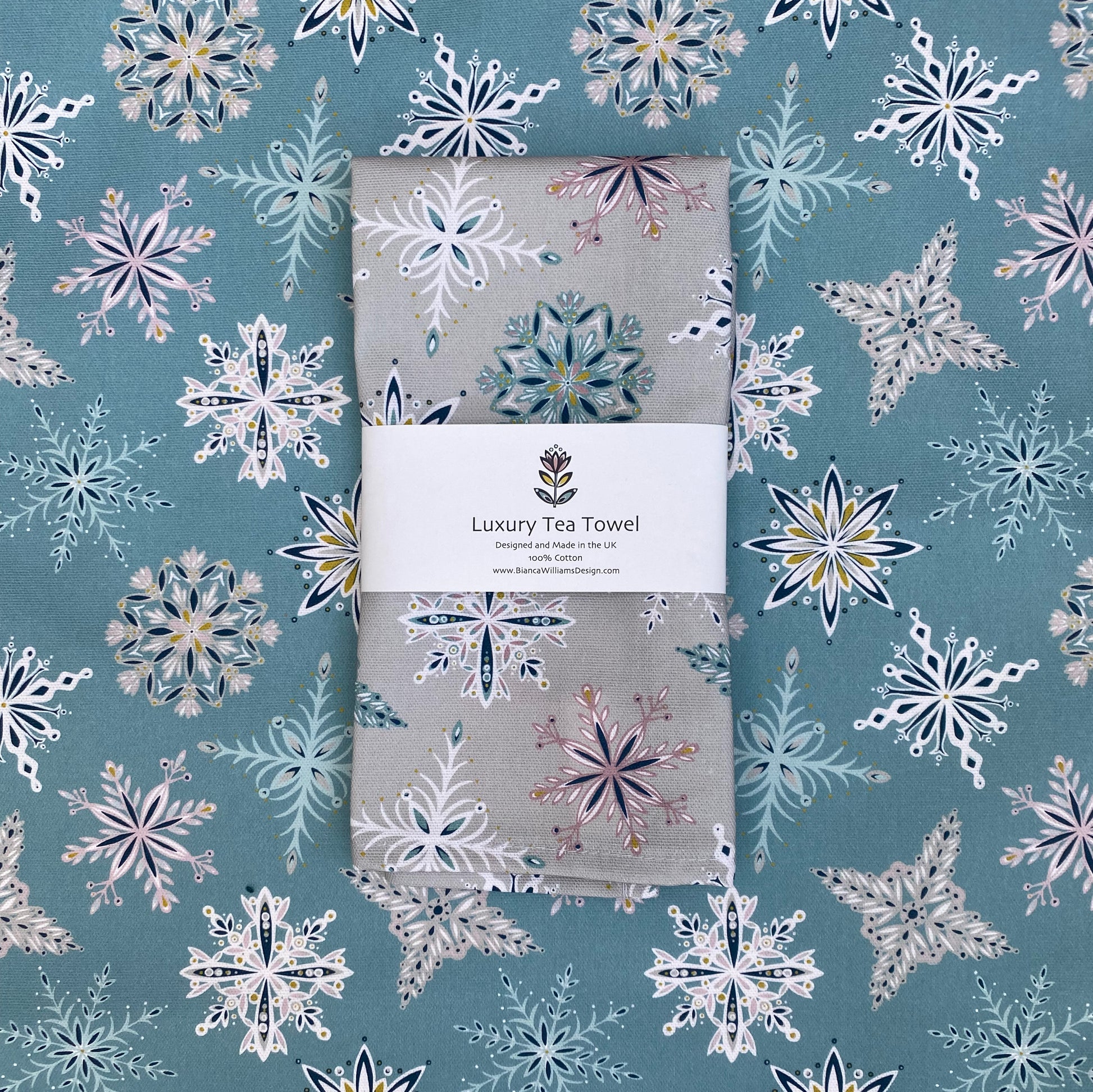 A silver grey snowflake tea towel has been packaged in a branded belly band and placed on an opened out tea towel featuring the same design but in a blue colourway.