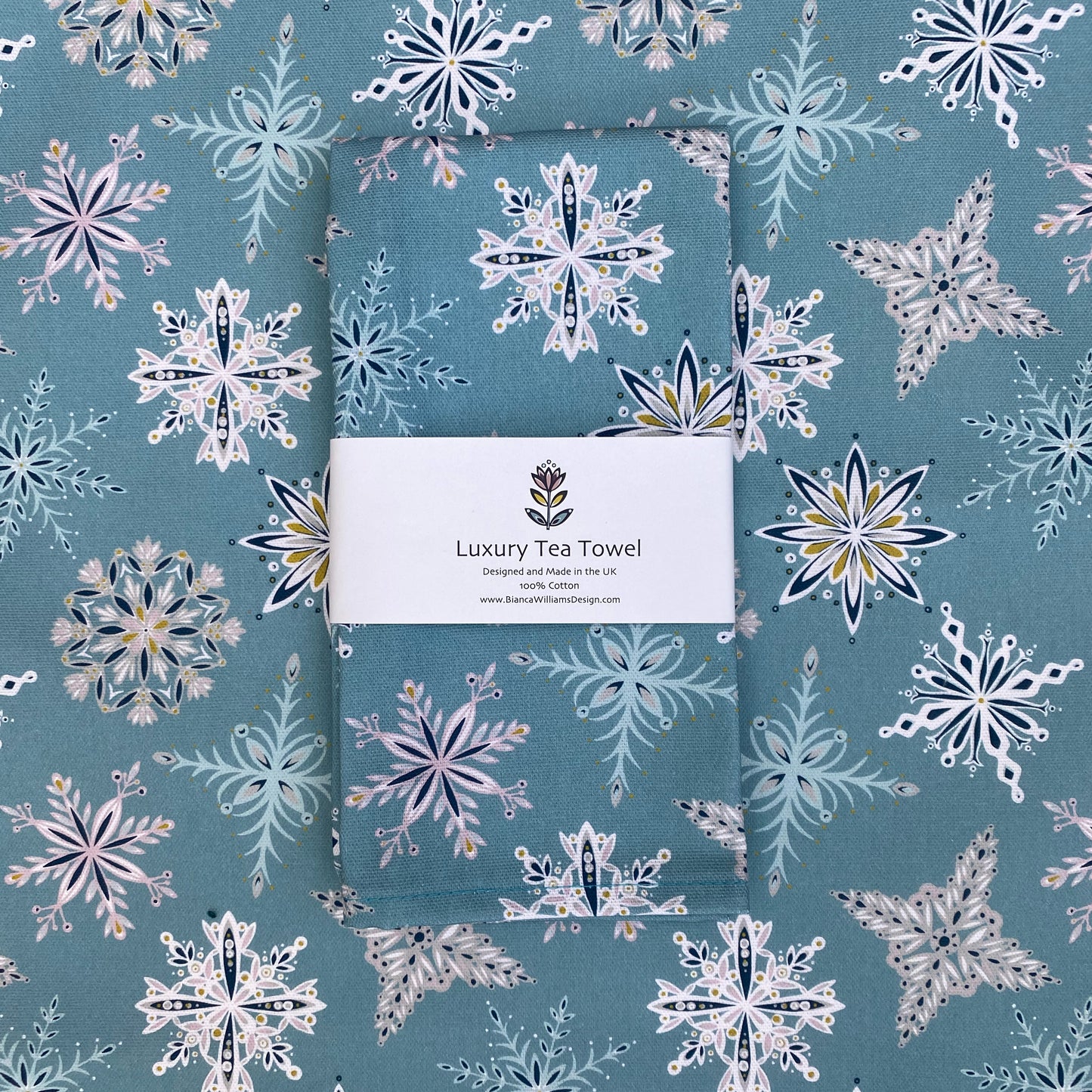 A Blue snowflake tea towel has been packaged in a branded belly band and placed on an opened out tea towel featuring the same design. The Snowflake design features white, teal, dusky pink and grey snowflakes on a blue background.